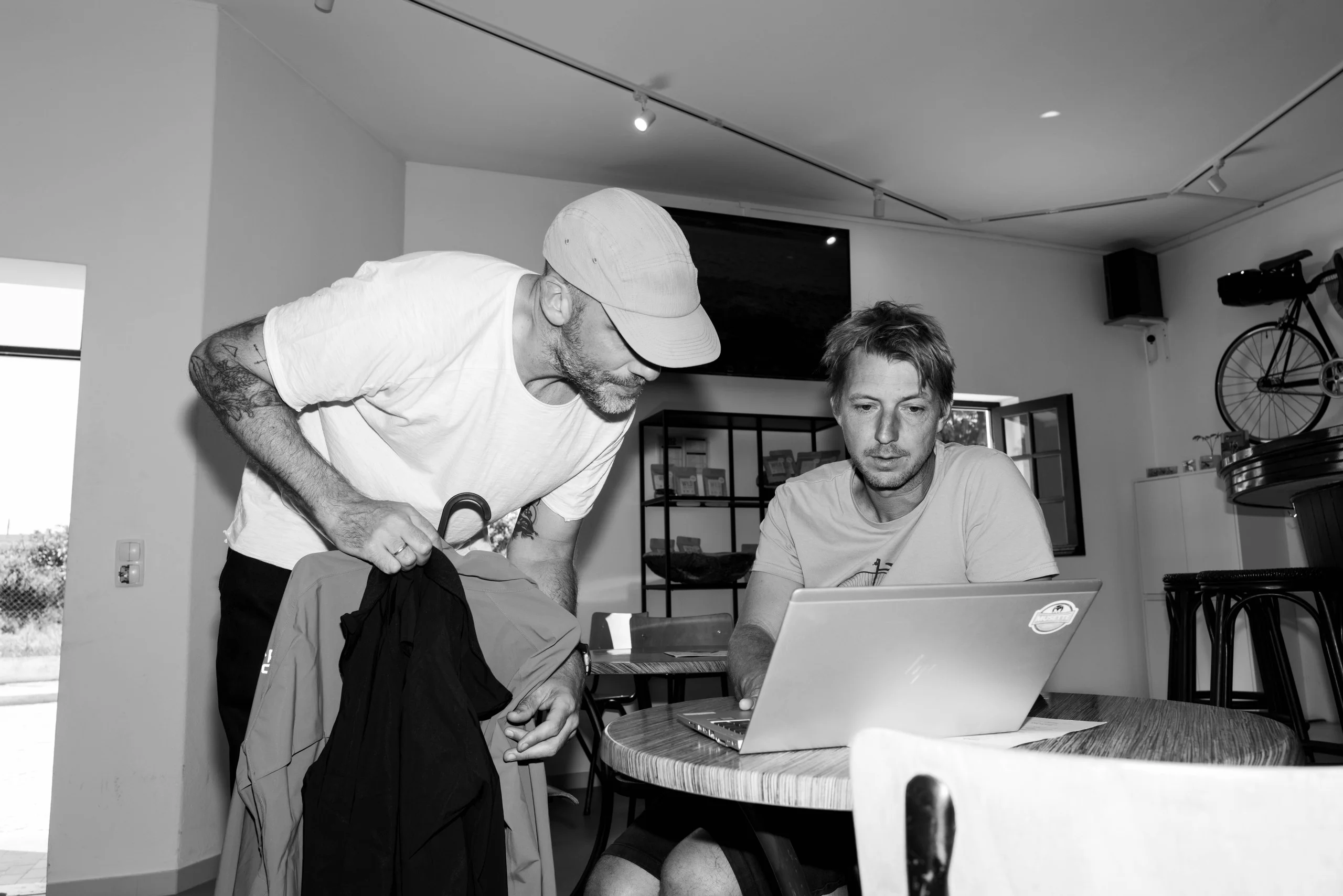 Jerom Pannier planning a cycling holiday on his laptop in the Musette café in Almádena, a rider unpacking beside him, in black and white
