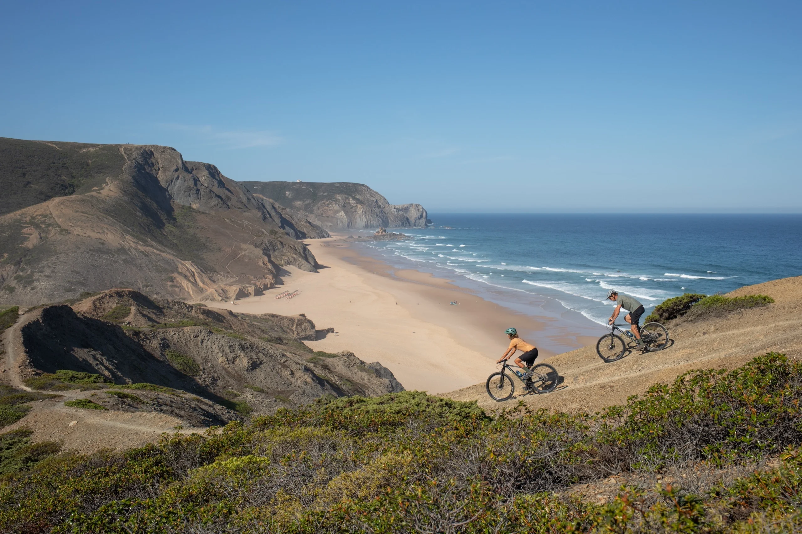 Two mountain bikers on a cliff path above a long empty beach on the Costa Vicentina, inside the natural park